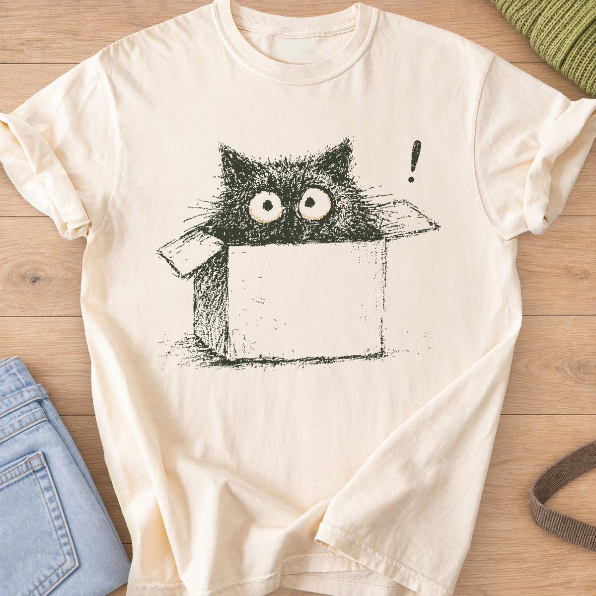 Wide-eyed black cat peeking out of a cardboard box beside an exclamation mark, dark charcoal ink on an Ivory T-Shirt