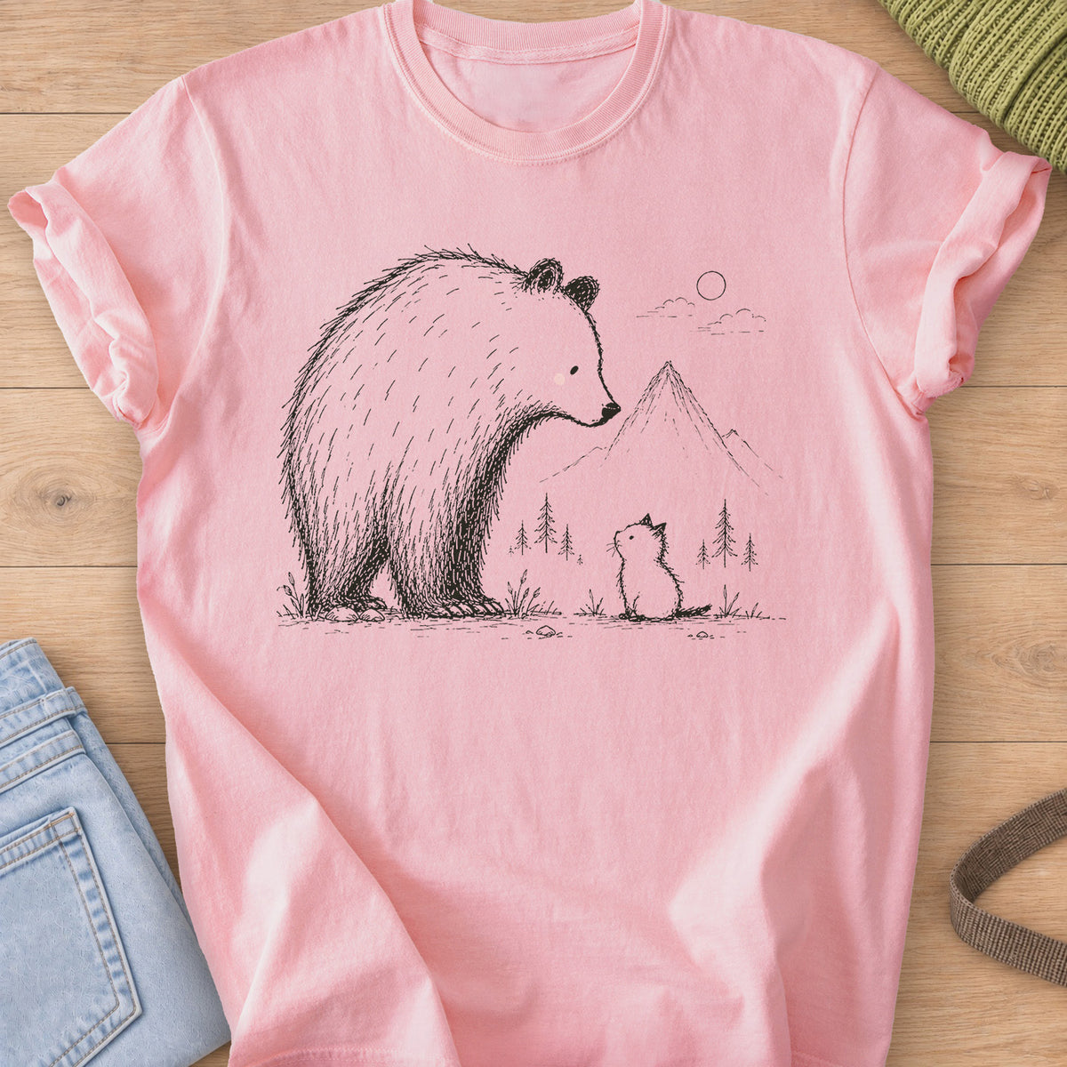 Small kitten sits looking up at a big bear, with mountains, pine trees and a sun, black ink on a Blossom T-Shirt