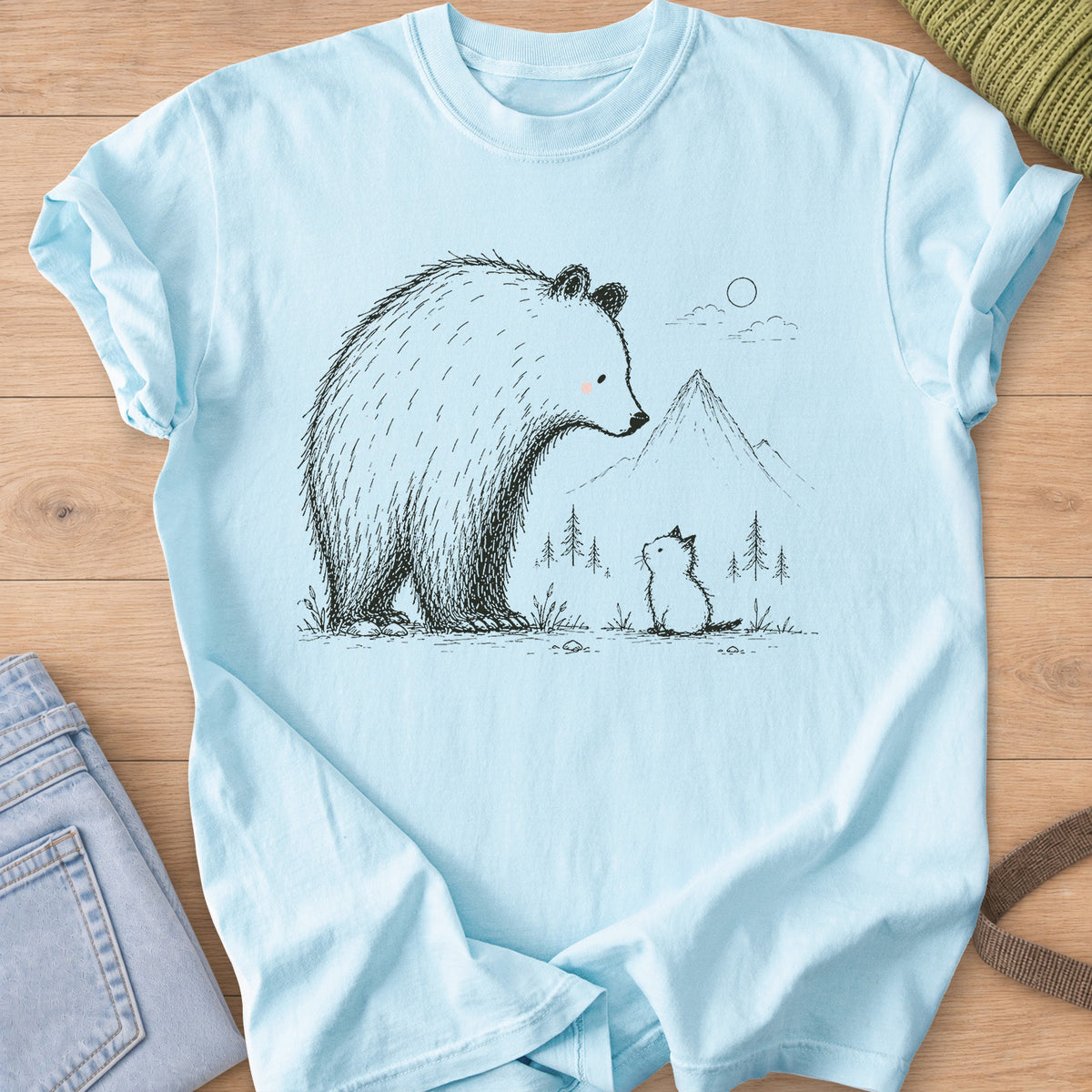 Small kitten sits looking up at a big bear, with mountains, pine trees and a sun, black ink on a Chambray T-Shirt