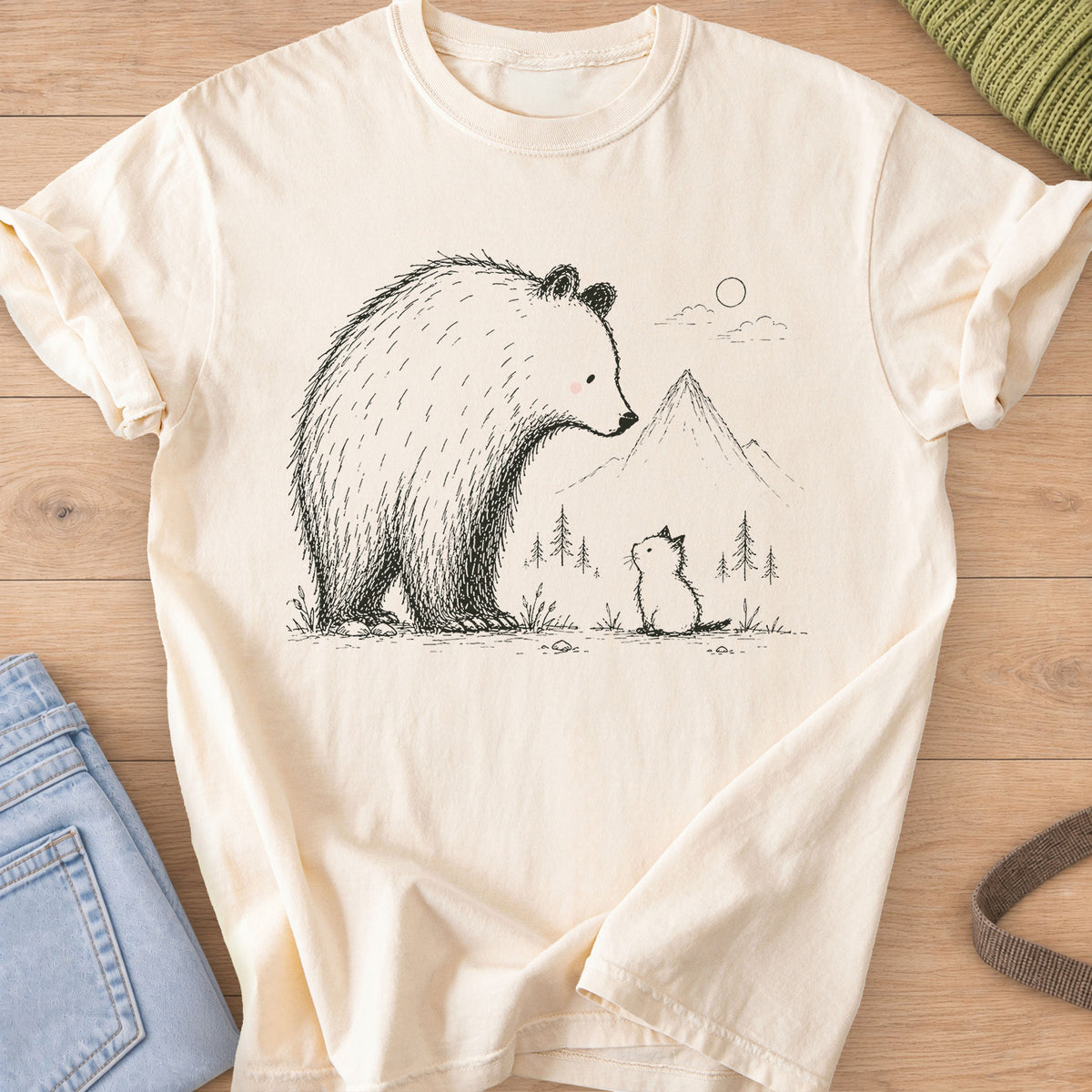 Small kitten sits looking up at a big bear, with mountains, pine trees and a sun, black ink on an Ivory T-Shirt