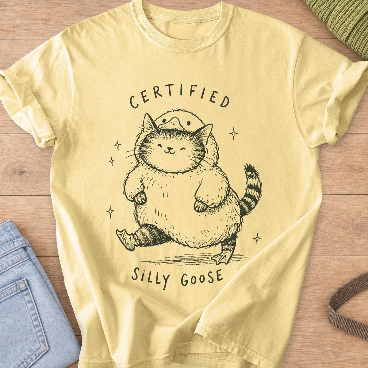 Chubby tabby cat in a goose costume walking among sparkles, "Certified Silly Goose", dark charcoal ink on a Butter T-Shirt