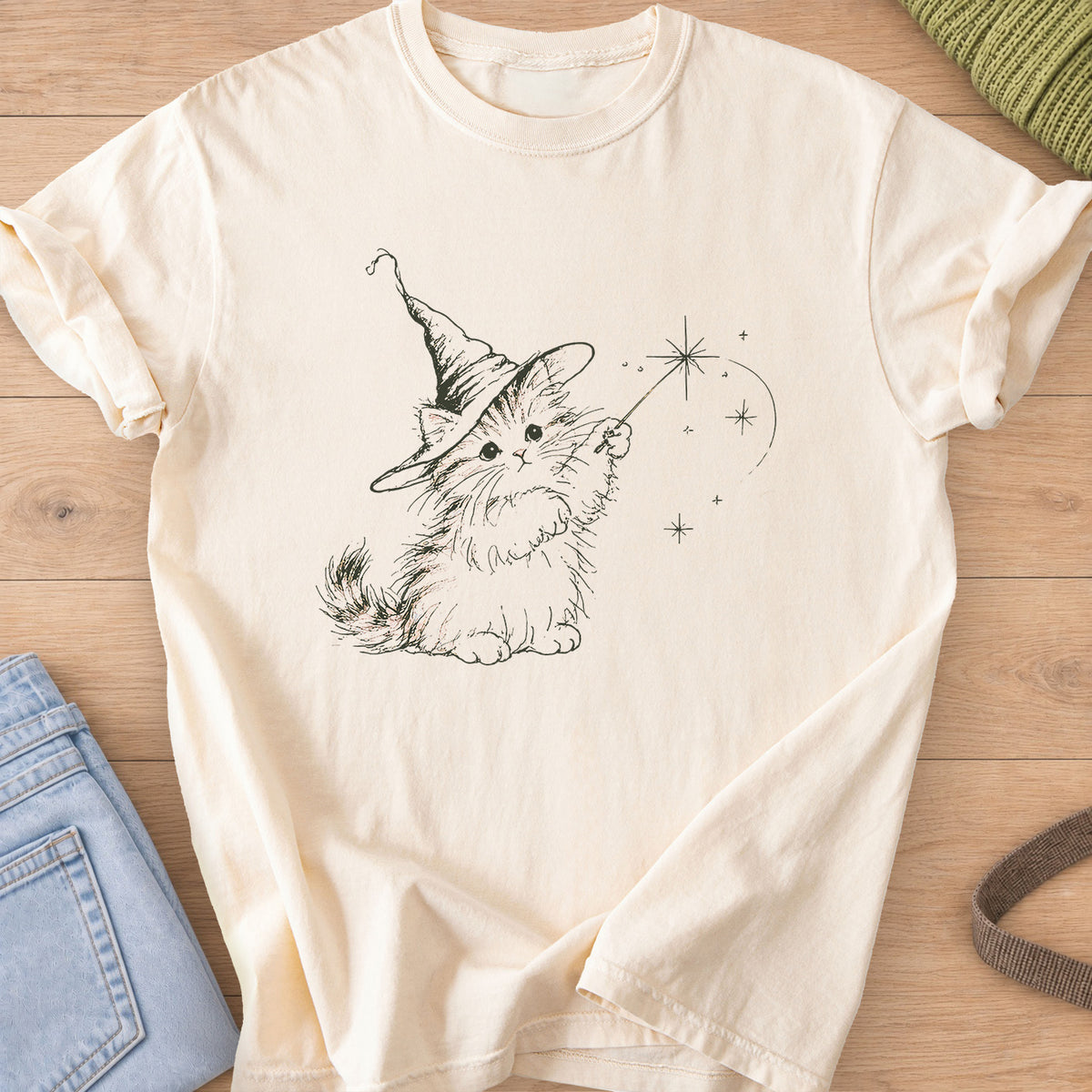 Fluffy kitten in a pointed wizard hat waving a magic wand with star sparkles, dark charcoal ink on an Ivory T-Shirt