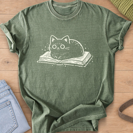 Round cat loaf with wide eyes resting on an open book, small dots around it, cream ink on a Moss T-Shirt
