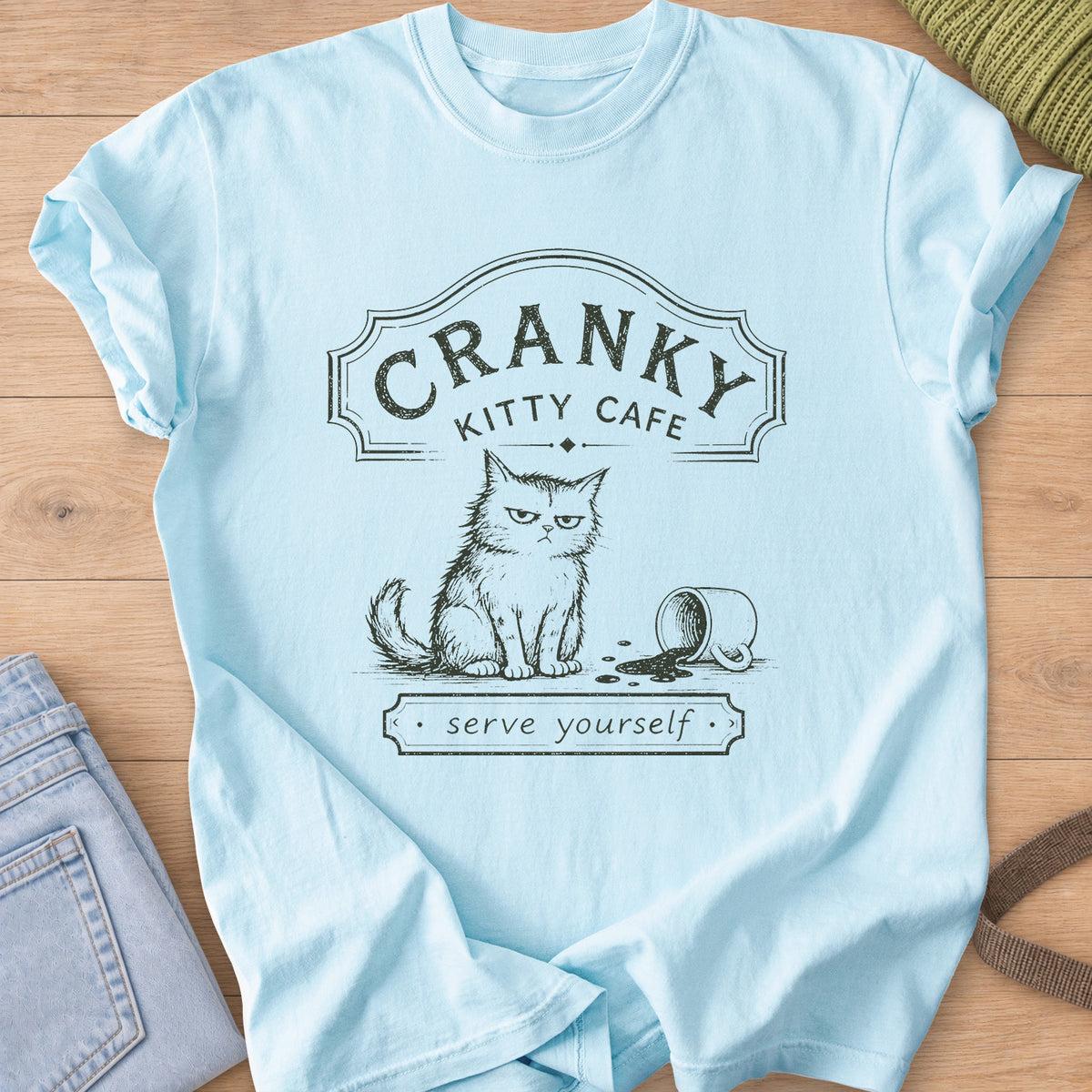 Grumpy fluffy cat beside a spilled coffee mug under a "Cranky Kitty Cafe" sign, dark charcoal ink on a Chambray T-Shirt