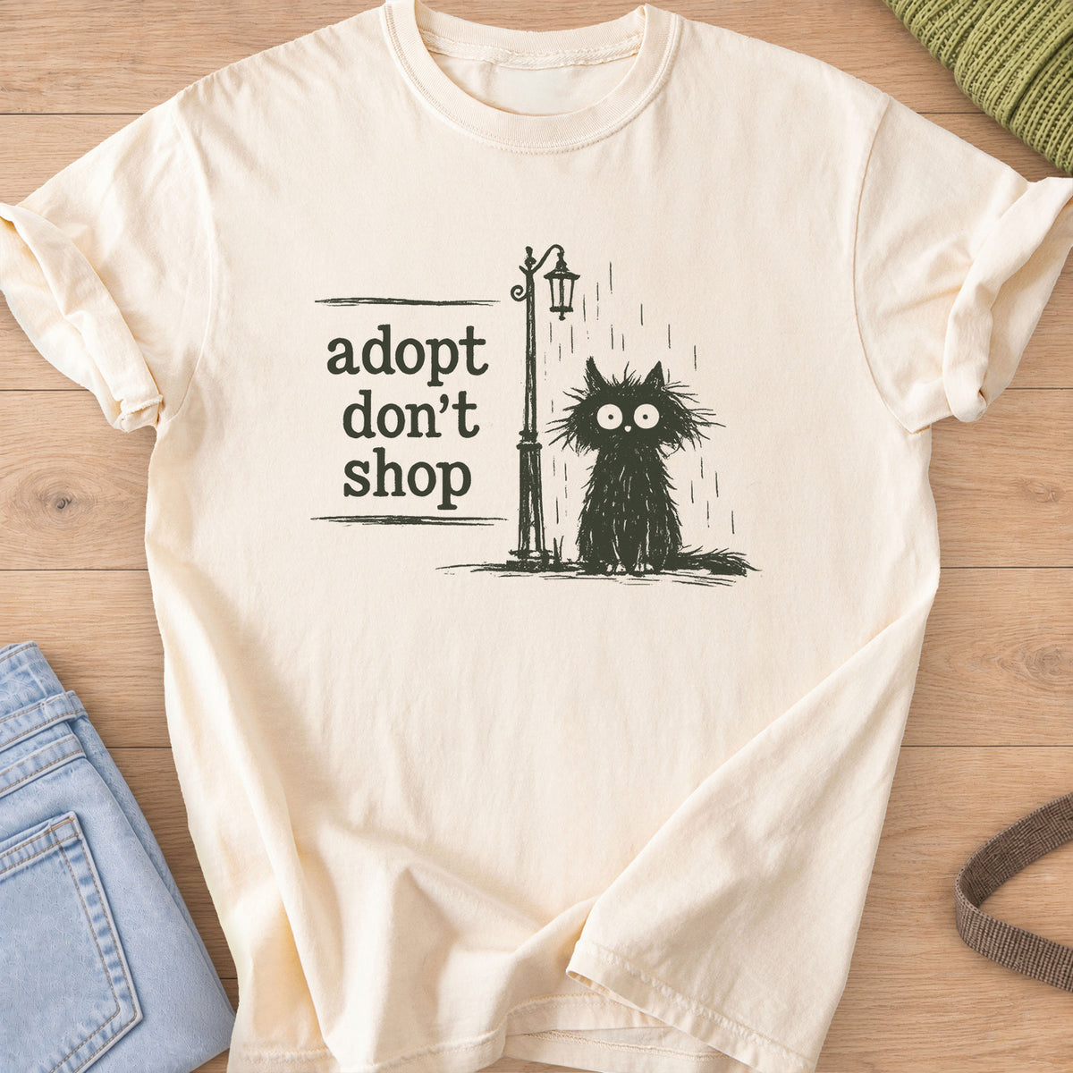 Scruffy black cat sitting in the rain by a street lamp, "adopt don't shop", dark charcoal ink on an Ivory T-Shirt