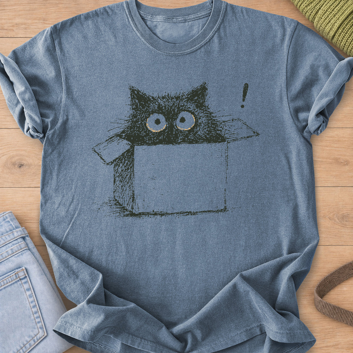 Wide-eyed black cat peeking out of a cardboard box beside an exclamation mark, dark charcoal ink on a Blue Jean T-Shirt