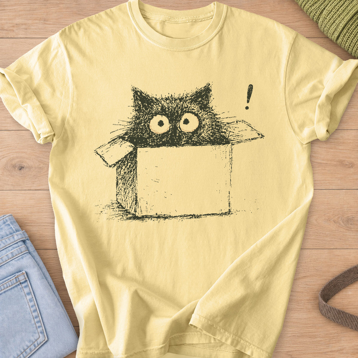 Wide-eyed black cat peeking out of a cardboard box beside an exclamation mark, dark charcoal ink on a Butter T-Shirt