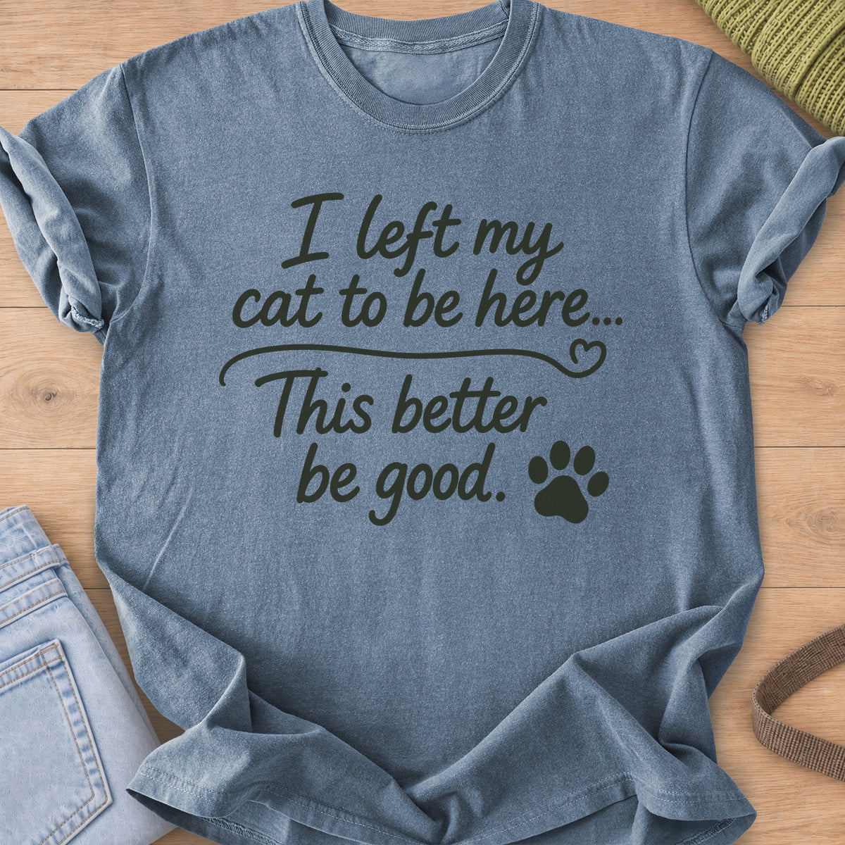 Script text "I left my cat to be here... This better be good." with a paw print, dark charcoal ink on a Blue Jean T-Shirt