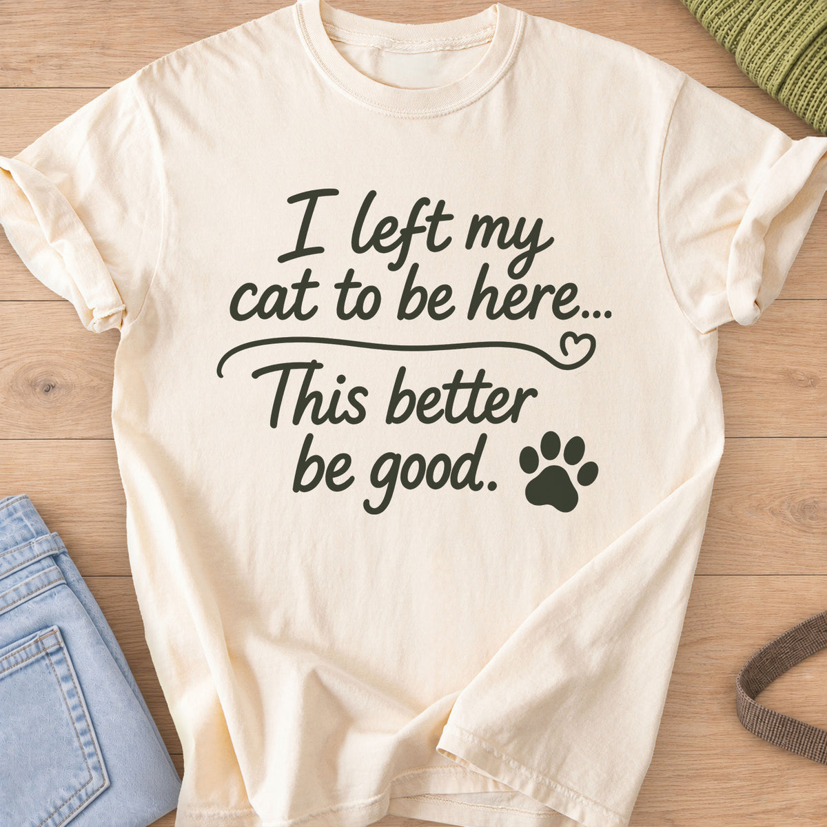 Script text "I left my cat to be here... This better be good." with a paw print, dark charcoal ink on an Ivory T-Shirt