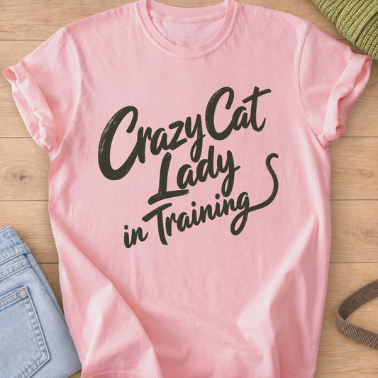 Script lettering reading "Crazy Cat Lady in Training" ending in a cat tail, dark charcoal ink on a Blossom T-Shirt