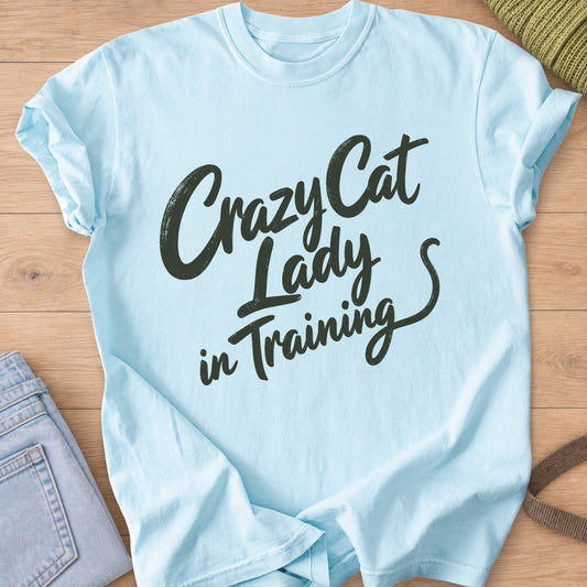 Script lettering reading "Crazy Cat Lady in Training" ending in a cat tail, dark charcoal ink on a Chambray T-Shirt