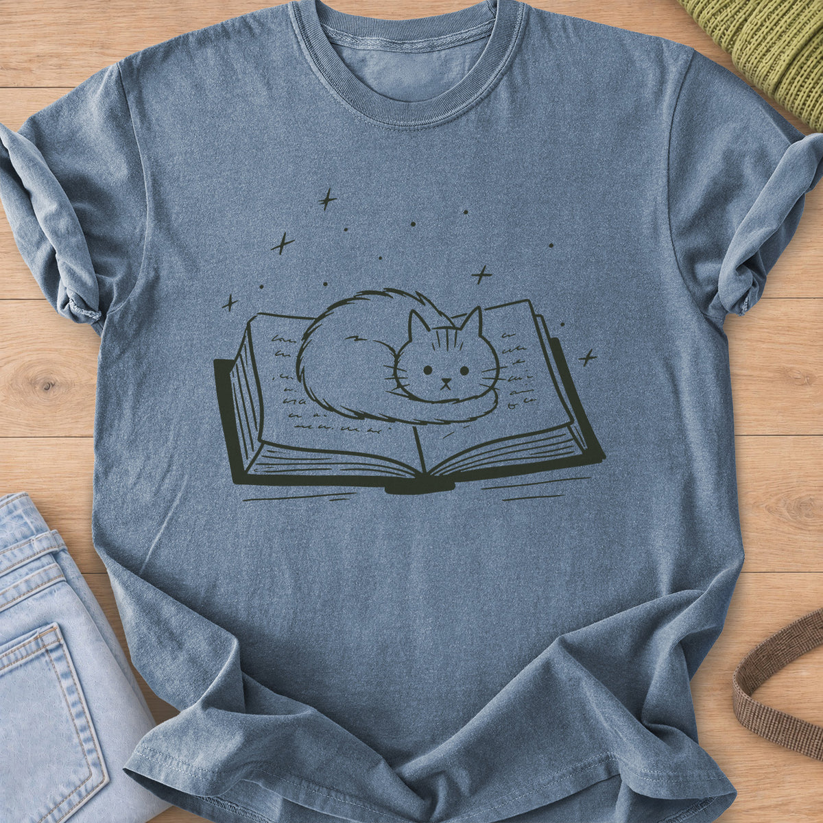 Fluffy cat curled up on an open book with sparkles and stars floating above, dark charcoal ink on a Blue Jean T-Shirt