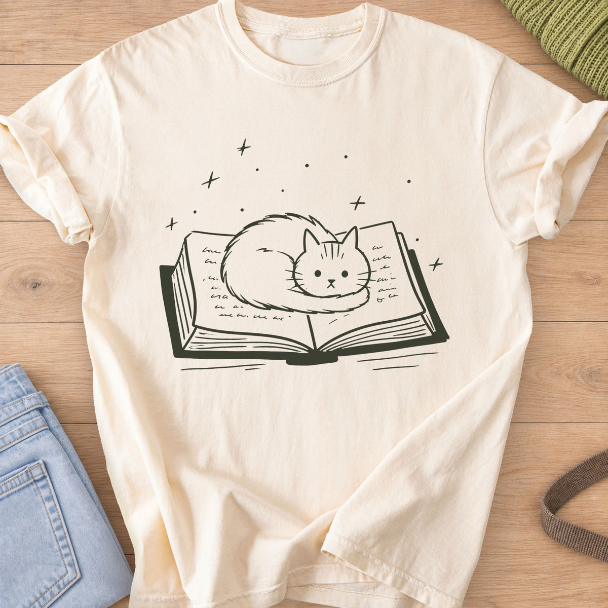 Fluffy cat curled up on an open book with sparkles and stars floating above, dark charcoal ink on an Ivory T-Shirt