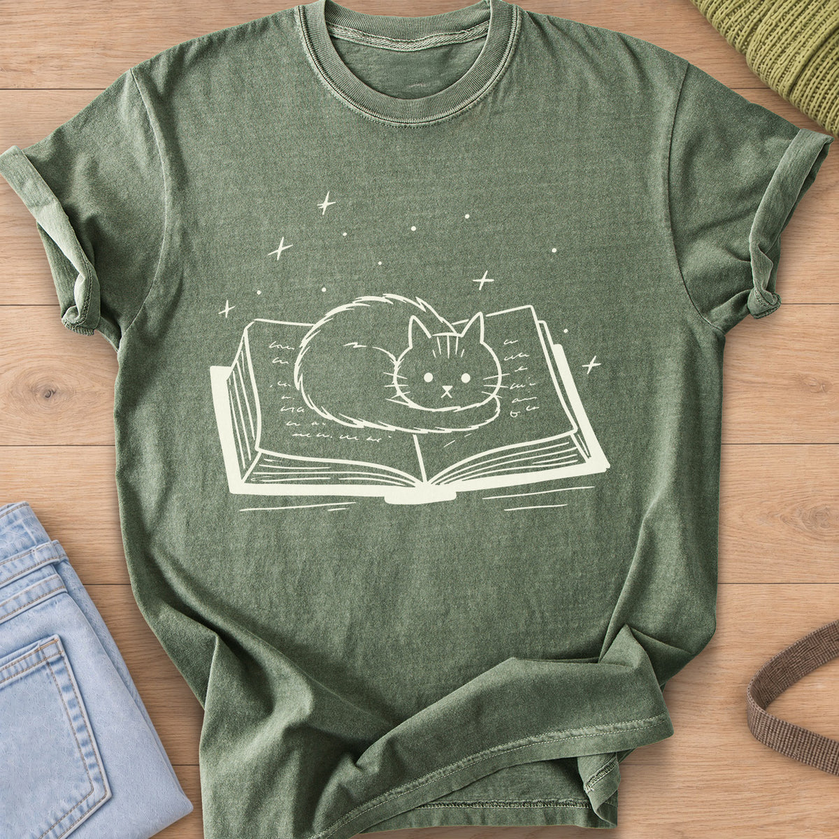 Fluffy cat curled up on an open book with sparkles and stars floating above, cream ink on a Moss T-Shirt