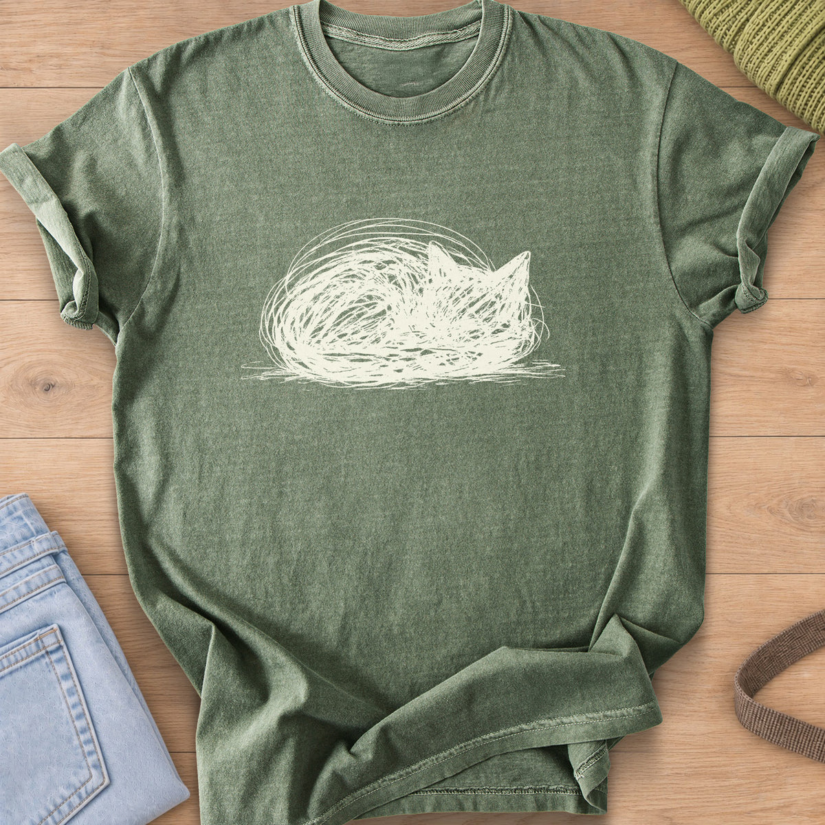 Scribbled cat curled into a round loaf with ears poking up, cream ink on a Moss T-Shirt