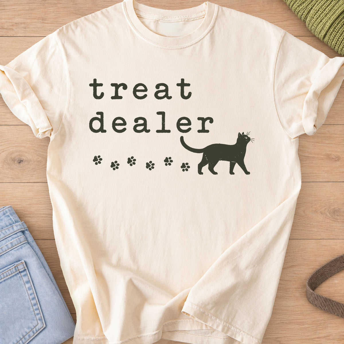 Walking cat silhouette with a trail of paw prints beneath the words "treat dealer", dark charcoal ink on an Ivory T-Shirt