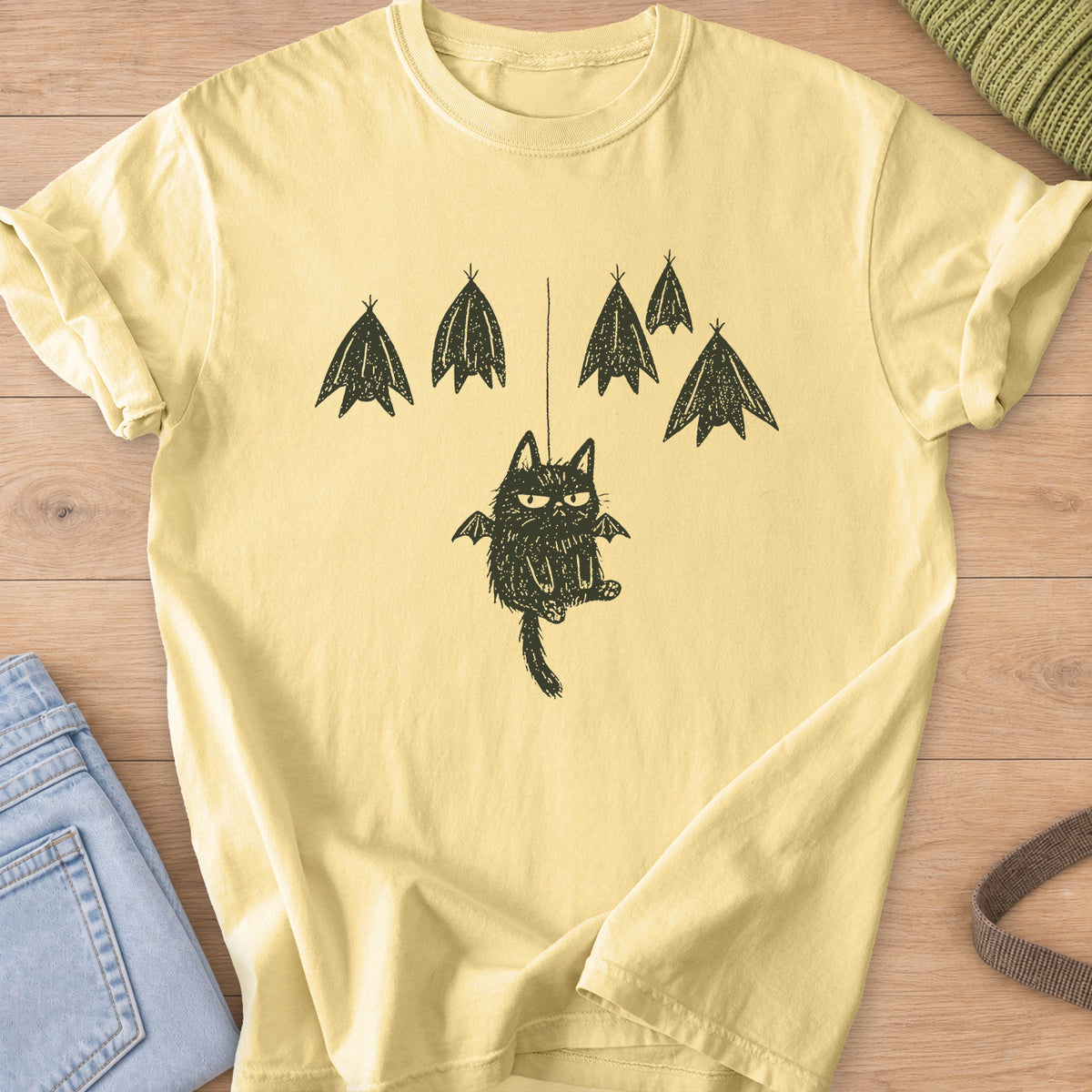 Grumpy fuzzy cat with bat wings dangling from a string among roosting bats, dark charcoal ink on a Butter T-Shirt