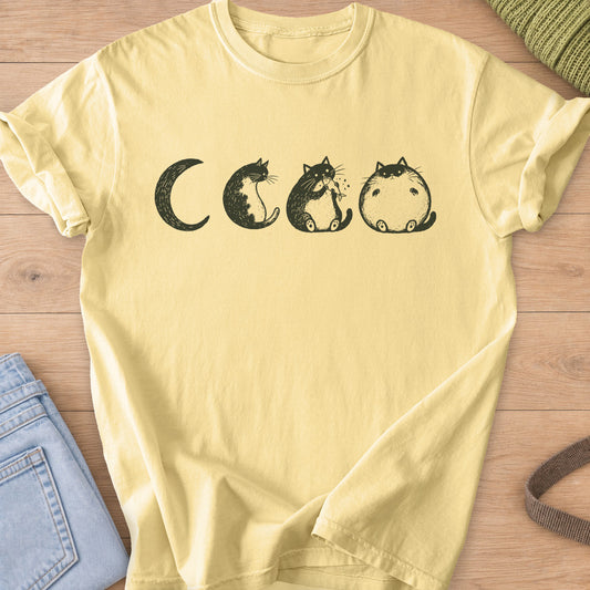 Four cats as moon phases, one eating a fish, ending in a round chubby full moon, dark charcoal ink on a Butter T-Shirt