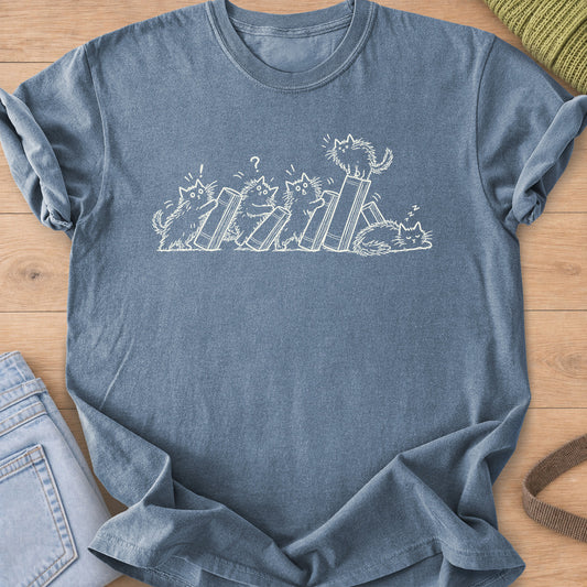 Five fluffy cats toppling books like dominoes, one perched on top, one asleep, cream ink on a Blue Jean T-Shirt