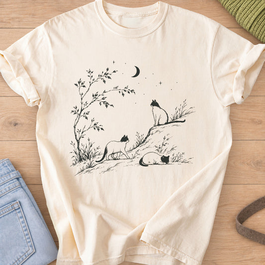 Three cats walking, sitting and sleeping on a hill by a tree under a crescent moon, dark charcoal ink on an Ivory T-Shirt