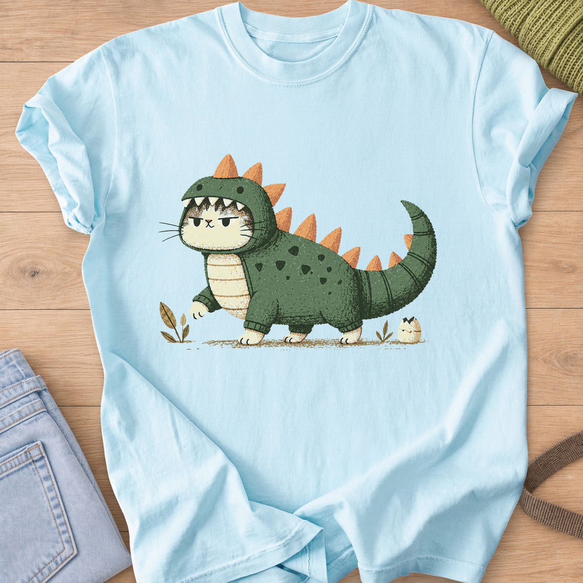 Grumpy cat in a spiky dinosaur costume walking past sprouts and a hatching egg on a Chambray T-Shirt