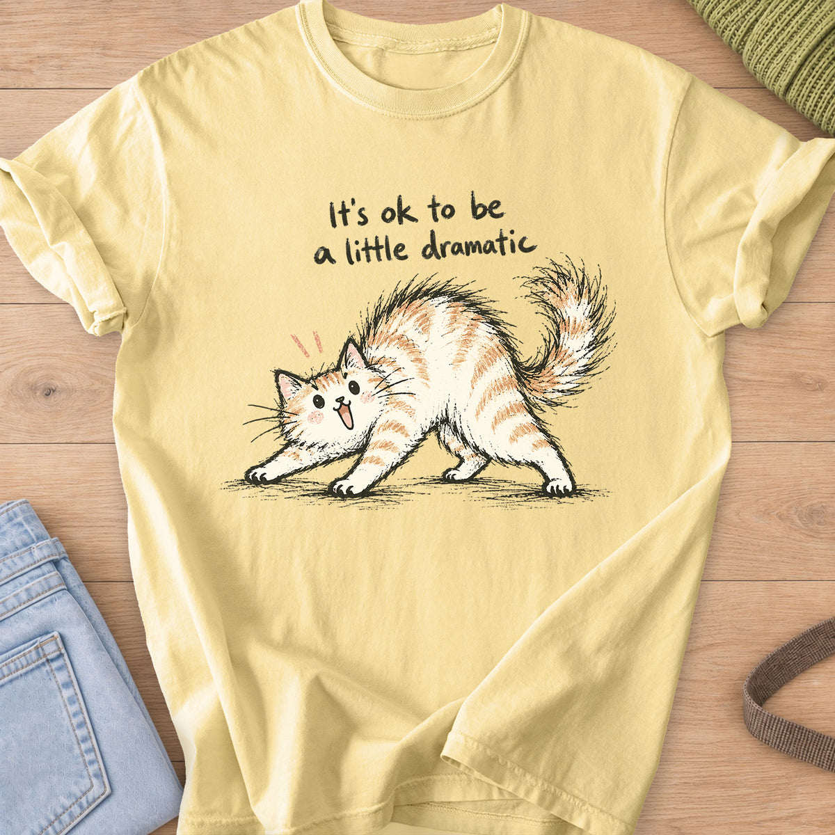 Startled fluffy cat arching its back beneath "It's ok to be a little dramatic" on a Butter T-Shirt