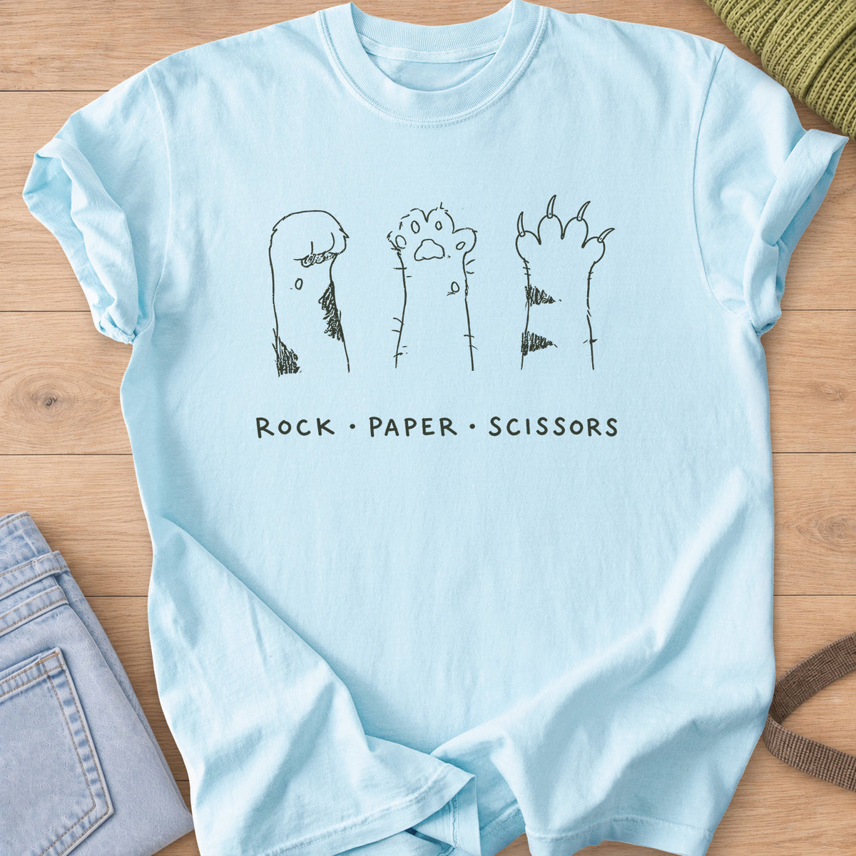 Three raised cat paws, curled, open and clawed, above "Rock Paper Scissors", dark charcoal ink on a Chambray T-Shirt