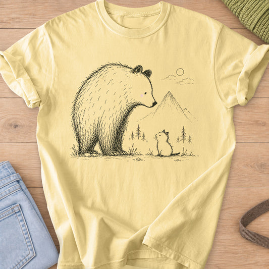 Small kitten sits looking up at a big bear, with mountains, pine trees and a sun, black ink on a Butter T-Shirt