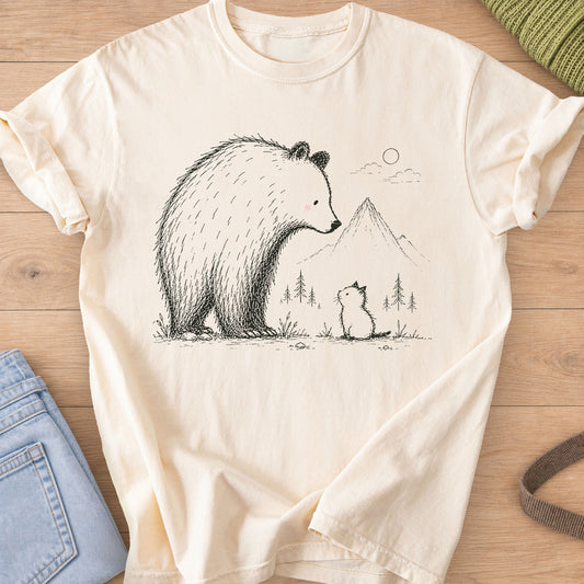 Small kitten sits looking up at a big bear, with mountains, pine trees and a sun, black ink on an Ivory T-Shirt