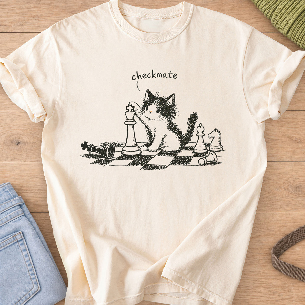 Fluffy kitten with a paw on a chess king, toppled pieces on a board, "checkmate", dark charcoal ink on an Ivory T-Shirt