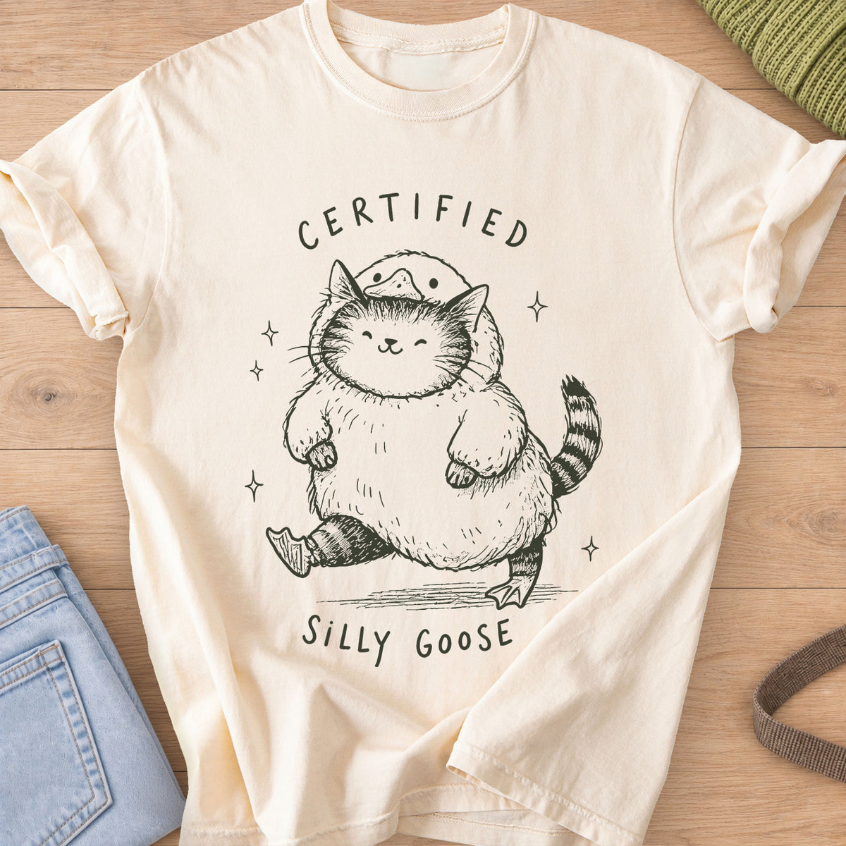 Chubby tabby cat in a goose costume walking among sparkles, "Certified Silly Goose", dark charcoal ink on an Ivory T-Shirt