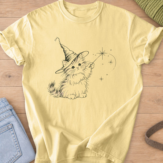 Fluffy kitten in a pointed wizard hat waving a magic wand with star sparkles, dark charcoal ink on a Butter T-Shirt