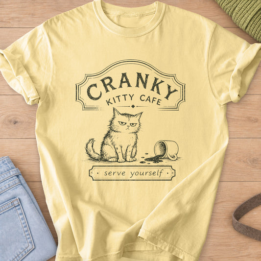 Grumpy fluffy cat beside a spilled coffee mug under a "Cranky Kitty Cafe" sign, dark charcoal ink on a Butter T-Shirt