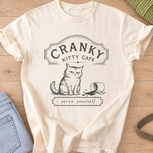 Grumpy fluffy cat beside a spilled coffee mug under a "Cranky Kitty Cafe" sign, dark charcoal ink on an Ivory T-Shirt